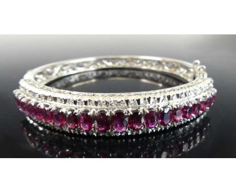 A white metal ruby and diamond hinged oval bangle, having 20 graduated oval cut rubies surrounded by 58 single cut diamonds, 