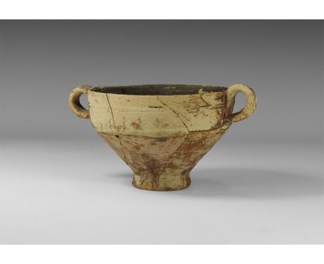 1000 BC.
A ceramic skyphos cup with pedestal foot, loop handles to the rim, indented groove around the rim, cream and brown s