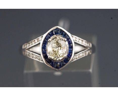 A white metal dress ring. Set with a central oval diamond of approximately 0.60cts. Surrounded by calibre cut sapphires and f