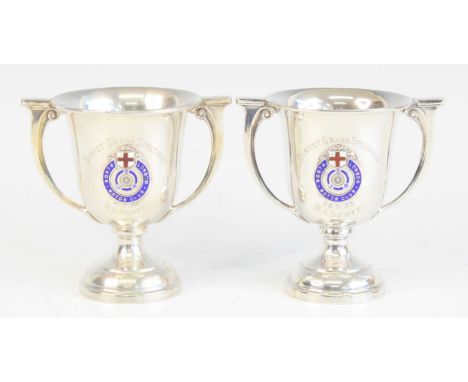 A near pair of George V. silver trophy cups, each having a wavy rim with cast border and twin handles, with enamelled badge f