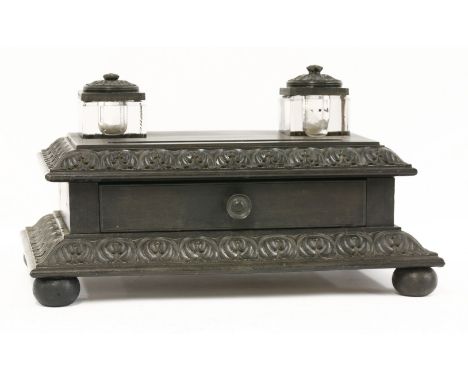An ebony desk stand,early 19th century, the two square glass inkwells with ebony covers, the base with a pen tray and frieze 