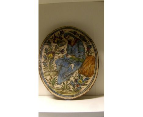 A 19th century Qajar pottery oval tile painted with a lady robed in blue seated on a rock amongst flowers, she holds a pen an