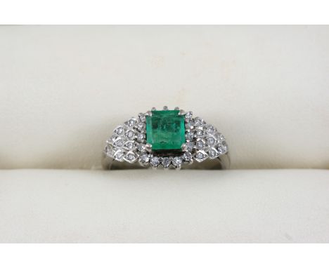 AN EMERALD AND DIAMOND CLUSTER RING the rectangular-shaped emerald is set within a surround of circular-cut diamonds, in whit