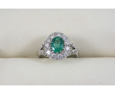 AN EMERALD AND DIAMOND CLUSTER RING the oval-shaped emerald is set within a surround of eleven circular-cut diamonds, in 18ct