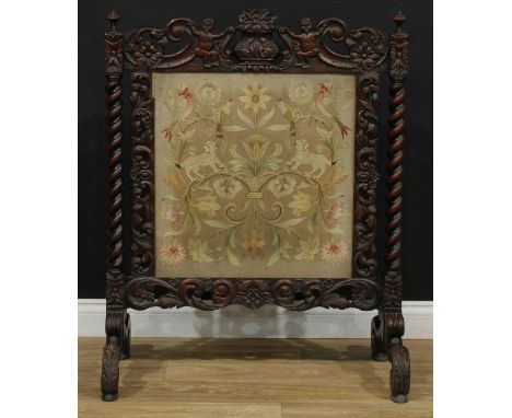 A Victorian oak cheval fire screen, shaped cresting carved with a flowering urn flanked by waving putti, the needlework banne