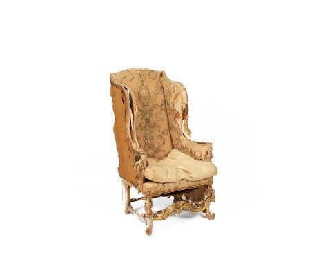 A Queen Anne wingback armchair in distressed velvet and embroidered foliate upholstery, on carved scrolling gilded legs and s