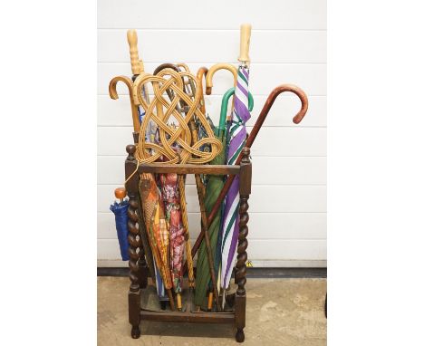 A large mixed collection of walking sticks, umbrellas and shooting stick contained within a wooden stick stand. 