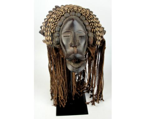 A Dan mask, Ivory Coast, the headpiece adorned with cowry shells and pods, with plaited fibre coiffeur, the mask height 38cm,