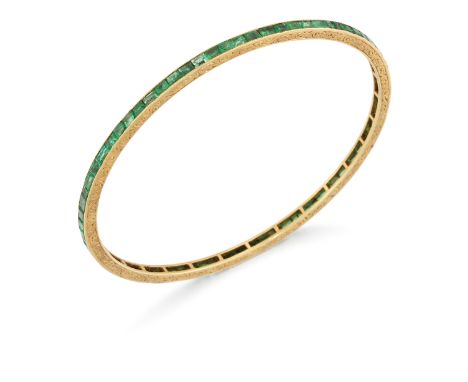 An early 20th century gold emerald bangle, a row of channel set calibré emeralds, to engraved guards, width 3mm, internal dia