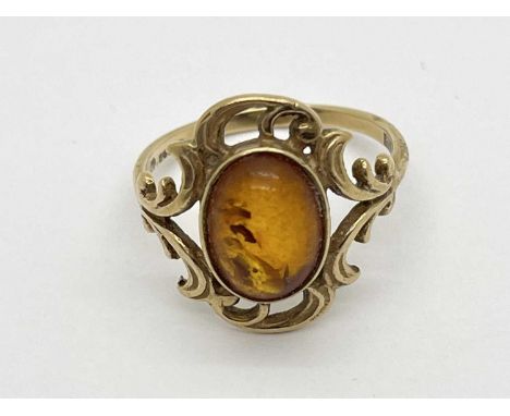 A 9ct Gold Dress Ring, the amber coloured cabochon stone collet set to centre, between highly decorative scroll setting (fing