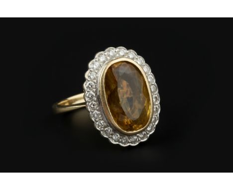 A TOPAZ AND DIAMOND CLUSTER RING, the oval mixed-cut topaz of brownish-yellow hue, collet set within a border of round brilli