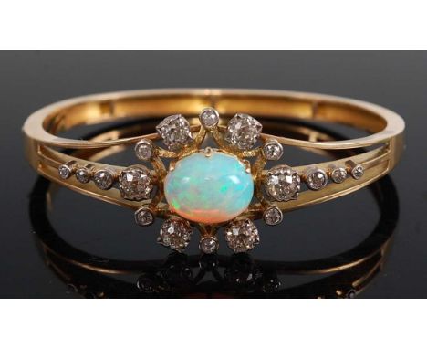 A yellow metal, opal and diamond hinged oval bangle, featuring a centre oval opal cabochon, having a pale body colour with bl