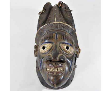 An African carved wooden and painted tribal mask, wearing a textured headdress, suspended with cowrie shells, hoops and a bel