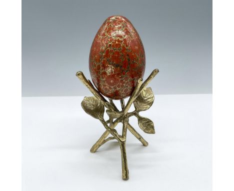 A beautiful hand-painted decorative egg decorated in gold, black, and orange standing on a leaf shaped tripod like display st