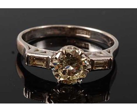 A white metal diamond solitaire ring, featuring a round brilliant cut diamond in a six claw setting, with a baguette cut diam