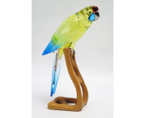 A Swarovski crystal parrot, mounted on a shaped wood Perch, 22cm high. A/F