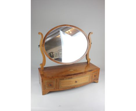 An oval mahogany inlaid toilet mirror on box base with bow front and three drawers, raised on bracket feet, 57cm