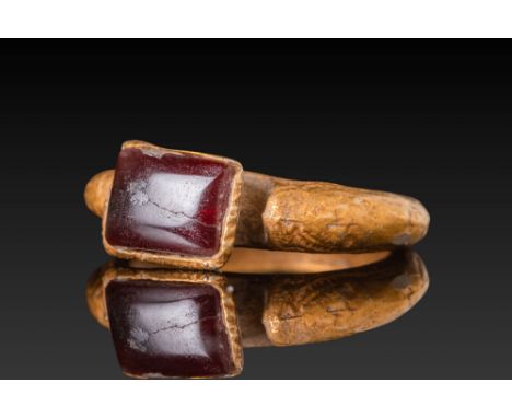 Ca. AD 400 - 600. A gold ring with a broad hoop and sharply angled shoulders. The projecting bezel is set with a deep red rec