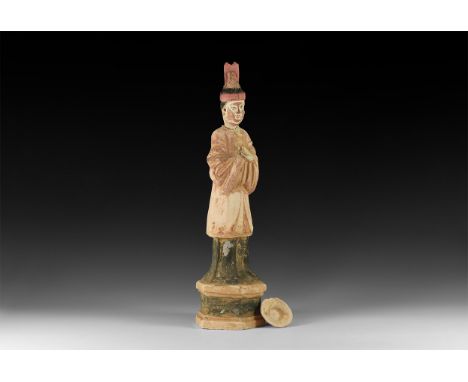 An undated archaistic ceramic statuette of a standing figure in loose smock and trousers in a hexagonal tiered base; hands cl