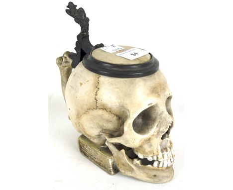 An unusual novelty porcelain tankard in the form of a human skull. The skull itself perched upon a book, with metal mounted l
