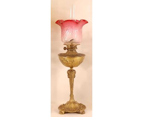 An Art Nouveau gilt metal pedestal oil lamp, having a cranberry tinted and plain acid etched moulded shade, over a naturalist