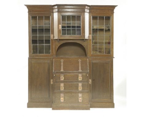 An oak secretaire bookcase,probably by Liberty & Co., with three leaded glass cupboards over an arched shelf, a fall front bu
