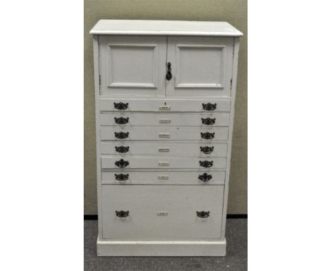 A vintage white painted tallboy with two doors, 6 short drawers and one deep drawer, drawers with nailed contents labels, 114