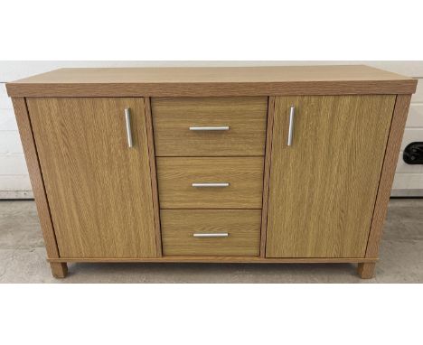 A modern light wood effect, sideboard with 2 cupboards and 3 central drawers. Long silver coloured handles, cupboards have in