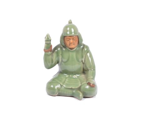 A Chinese celadon glazed stoneware model of a warrior, sitting cross-legged holding a lamp, impressed character marks to base