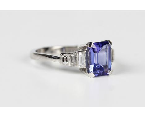 A platinum, tanzanite and diamond ring, claw set with the cut cornered rectangular step cut tanzanite between baguette cut th