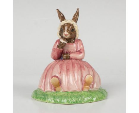 A figure of a bunny wearing a pink dress and white bonnet, sitting on the grass and holding a flower. Bunnykins of the Year 2