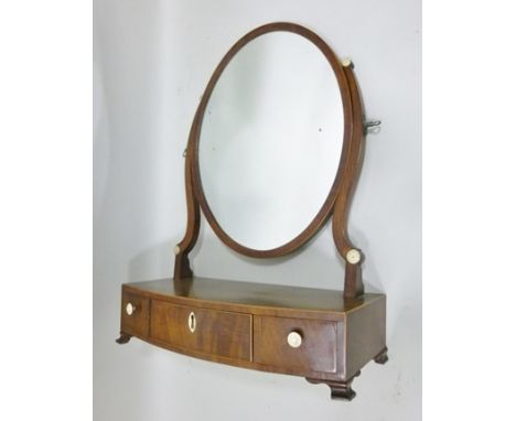 A 19TH CENTURY MAHOGANY TOILET MIRROR having oval swing plate held between twin shaped horns, mounted on a bow fronted box ba