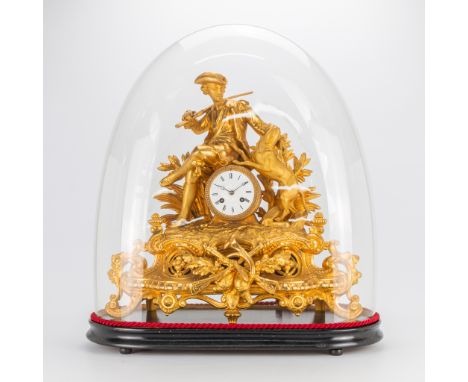 A mantle clock, with a hunter figurine and his dog, hunting trophies. Under a glass dome. 19th century. (25 x 56 x 62 cm)