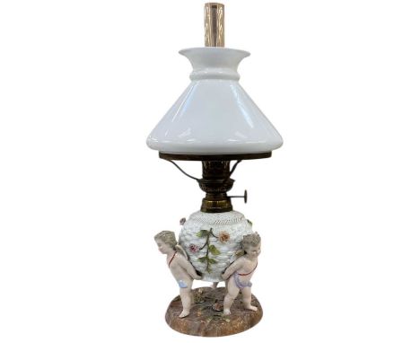 An early 20th century Continental porcelain oil lamp, modelled with the well held aloft by three cherubs, with opaque glass s