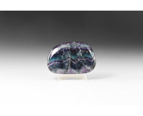 An oval polished specimen of fluorite, with dark purple and green colour zigzag pattern; natural fractures to the body with '