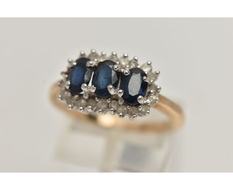 A 9CT GOLD SAPPHIRE AND DIAMOND CLUSTER RING, desinged with three oval cut deep blue sapphires, each claw set, within a surro