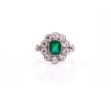 An Edwardian emerald and diamond cluster ring; the stepped rounded rectangular emerald in milegrain mount with a border of te