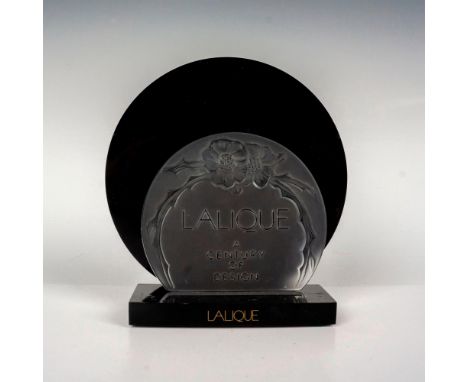A round clear plaque that reads Lalique - A Century of Design with a black base. Plaque signed Lalique France. Dimensions of 