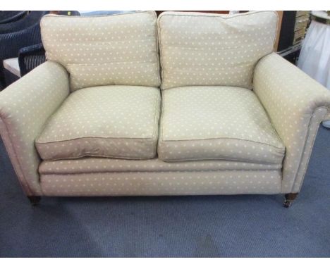 A modern two seater sofa in a beige fabric with a cream leaf design, above mahogany feet with castors, manufactured by The Od