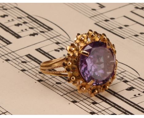 A tested 18ct gold synthetic pinkish-purple colour change sapphire round brilliant cut solitaire ring, with floral design col