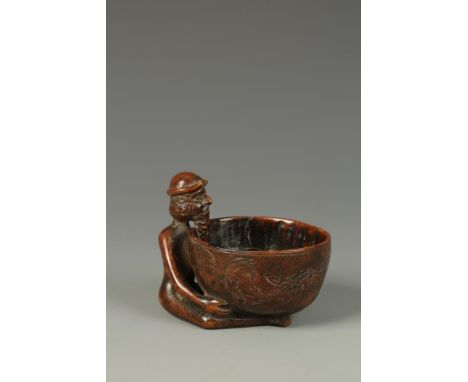 A TREEN CUP in the form of a naked man sat legs apart holding a broad bowl, with all over carved decoration, probably South G