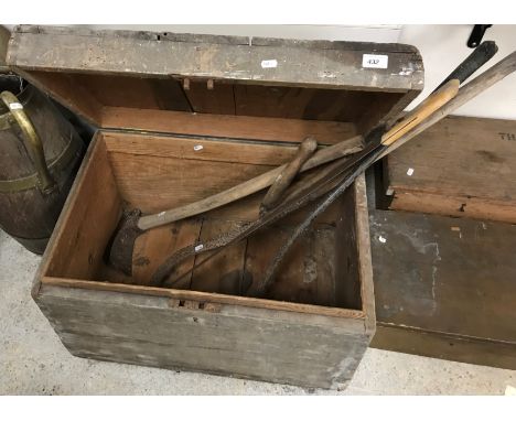 A wooden trunk containing various vintage hand tools comprising an adze, two scythes, a saw and a pair of shears 