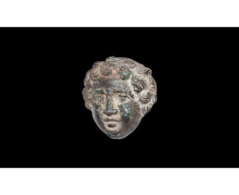 1st-2nd century AD. A bronze applique in the form of the face of Eros with finely detailed youthful features and bands of cur