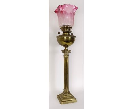 A brass oil lamp with a ruby tinted glass shade, the base in the form of a Corinthian column, 83 cm highsome small losses to 