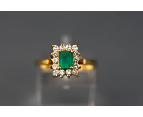 A modern yellow gold cluster ring set with a rectangular emerald and fourteen round brilliant diamonds. Stamped 18K. Size M. 