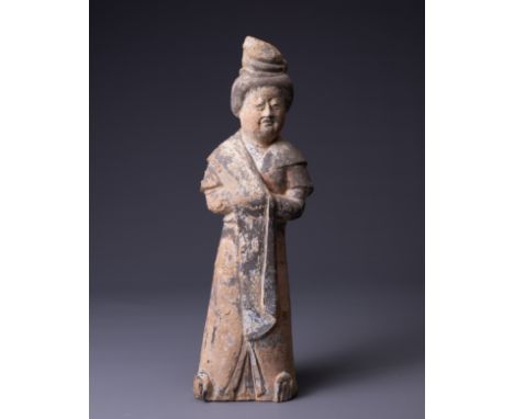 A CHINESE PAINTED GREY CARVED POTTERY FIGURE OF AN ATTENDANT, EARLY TANG DYNASTY (AD 618-907). The solid figure modelled stan