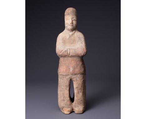 A CHINESE PAINTED POTTERY FIGURE OF AN ATTENDANT, TANG DYNASTY (AD 618-907). Modelled standing with his hands clasped to his 
