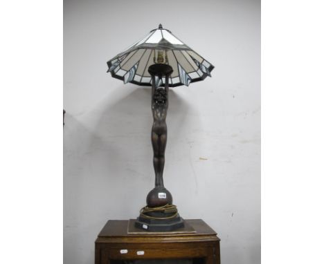 A Bronzed Effect Art Deco Style Table Lamp, with a leaded glass shade, female nude standing on a ball, stepped base.