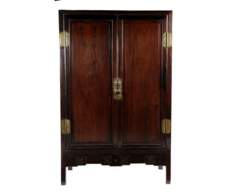 A Chinese hardwood cupboard, with brass mounts and a pair of panelled doors enclosing shelves and a pair of drawers, above a 