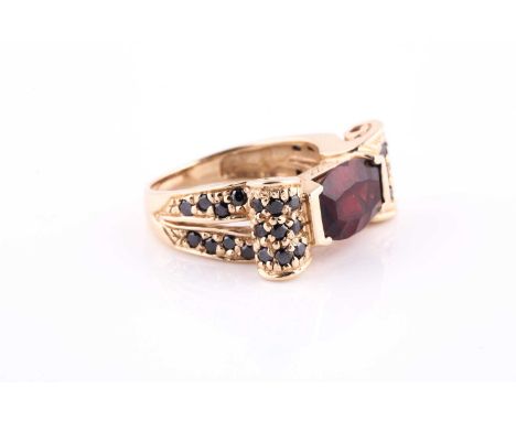 A 14k yellow gold, garnet, and black diamond ring, set east to west with a mixed fancy-cut garnet, the scrolled shoulders pav